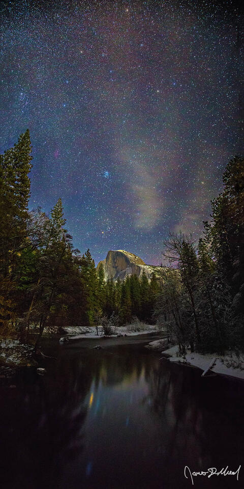 The stars shine brightly in the sky above Half Dome and the snow covered banks of Yosemite Valley as Half Dome reflects in the still waters of the Merced River