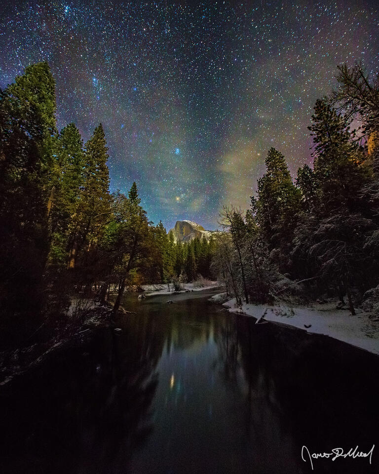 Stars shine brightly in the night sky above snow covered Yosemite Valley as Half Dome reflects in the still waters of the Merced River.