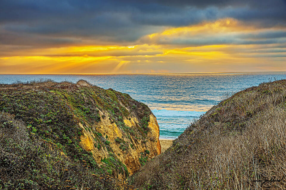 Turquoise water of the Pacific Ocean rolls ashore below the headlands at sunset as light rays shine through the clouds.
