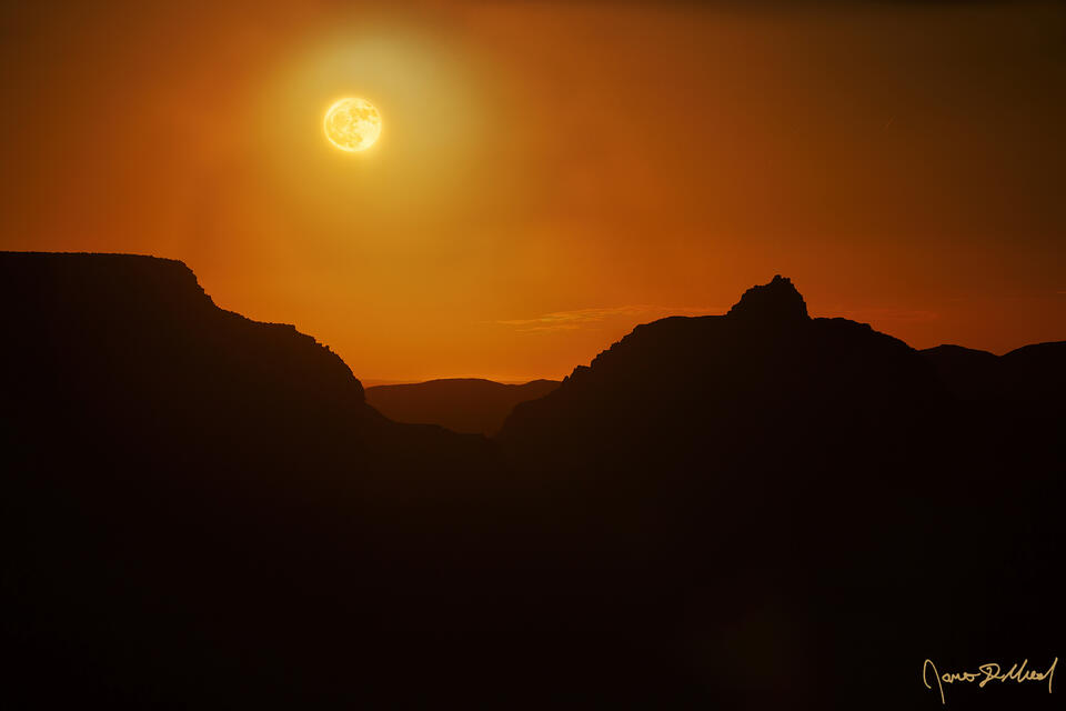 The full moon rises between Wotans Throne and Vishnu Temple in the Grand Canyon, lighting the night sky around it a brilliant orange as the foreground darkens
