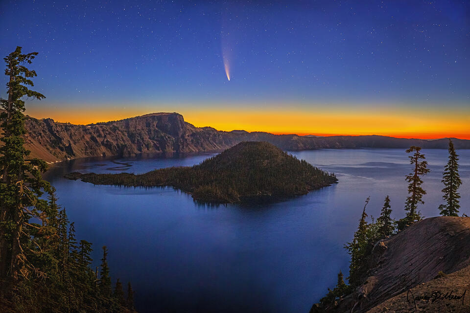 Comet NEOWISE hangs in a starry sky above Wizard Island at nautical dawn, as moonlight illuminates Llao Rock, the caldera rim, and the waters of Crater Lake