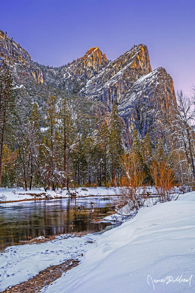 The setting sun illuminates the top of the Three Brothers high above the Merced River as snow blankets Yosemite Valley in the winter.