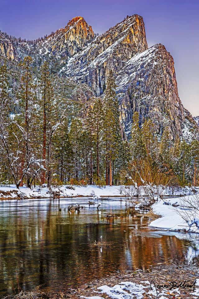 Sunset in the winter lights the top of the Three Brother as trees reflect in the still waters of the Merced River while snow blankets Yosemite Valley.