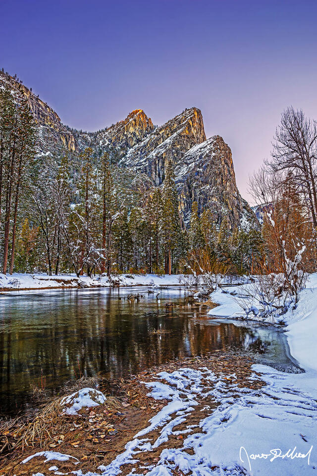 The setting sun illuminates the top of the Three Brothers high above the Merced River as snow blankets Yosemite Valley in the winter.
