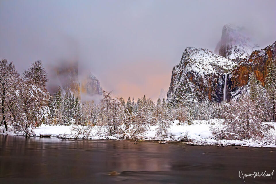 Fog envelope El Capitan and Cathedral Rocks as the rising moon behind the clouds illuminate the horizon with a warm glow while snow blankets Yosemite Valley