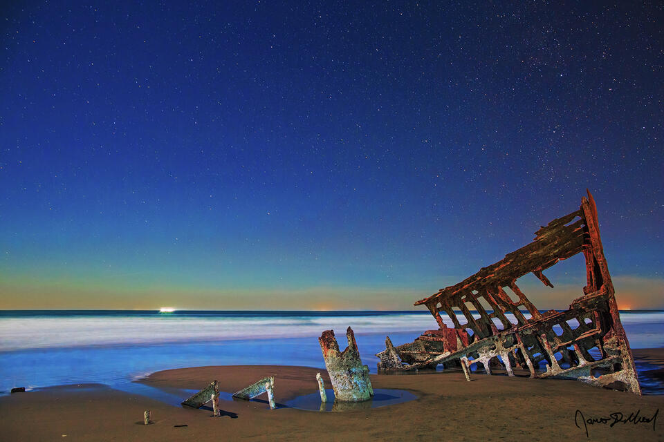 A Waxing Gibbous moon illuminates the Pacific Ocean and the Wreck of the Peter Iredale at night along the Oregon Coast.