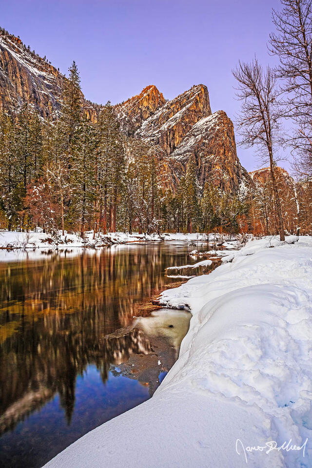 Alpenglow above the Three Brothers and the Merced River during twilight. Ice begins to form along the edges of the Merced River as snow blankets Yosemite Valley