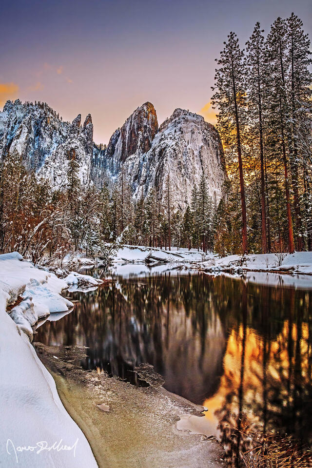 Sunset lights the skies overhead as Cathedral Rocks and Cathedral Spires reflect in a calm, partially frozen Merced River in snowy Yosemite Valley.