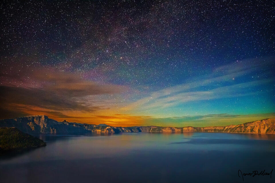 The Milky Way spiral arm rises above Crater Lake and a strong diffuse aurora as moonlight illuminates Wizard Island, Llao Rock, and the volcanic caldera rim.