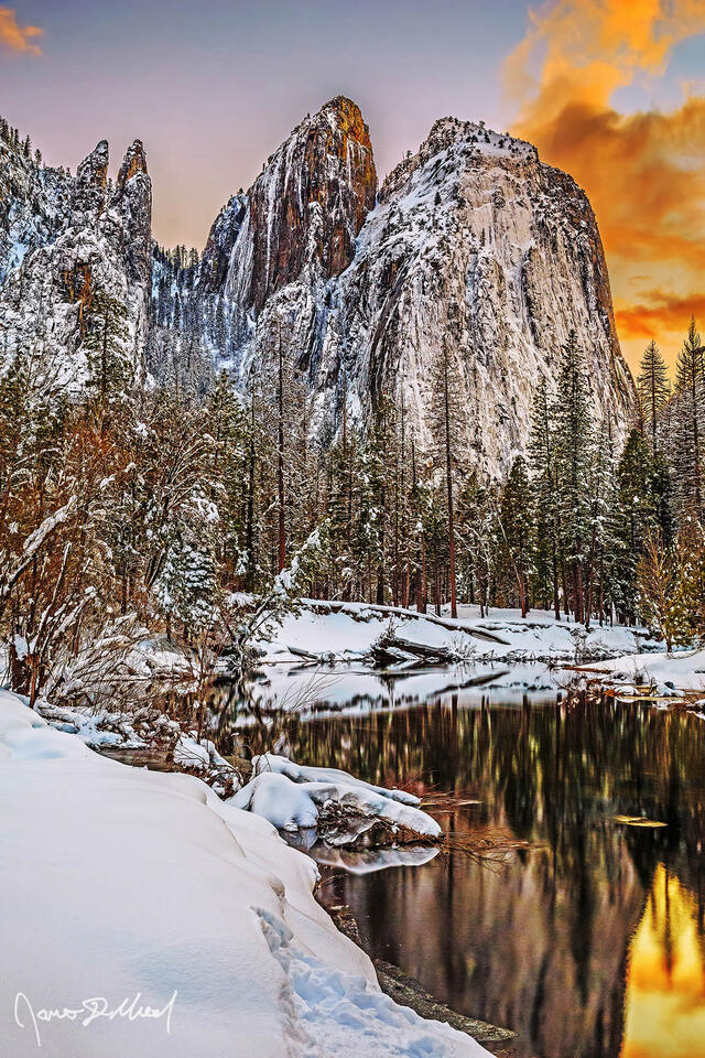 Sunset lights the clouds and skies overhead as Cathedral Rocks and Cathedral Spires reflect in a calm, partially frozen Merced River in snowy Yosemite Valley.