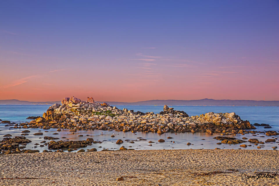 Dusk brings colors to the skies over the Pacific Ocean as civil twilight fades. Seagulls use a rocky outcrop just offshore for nesting and rest.