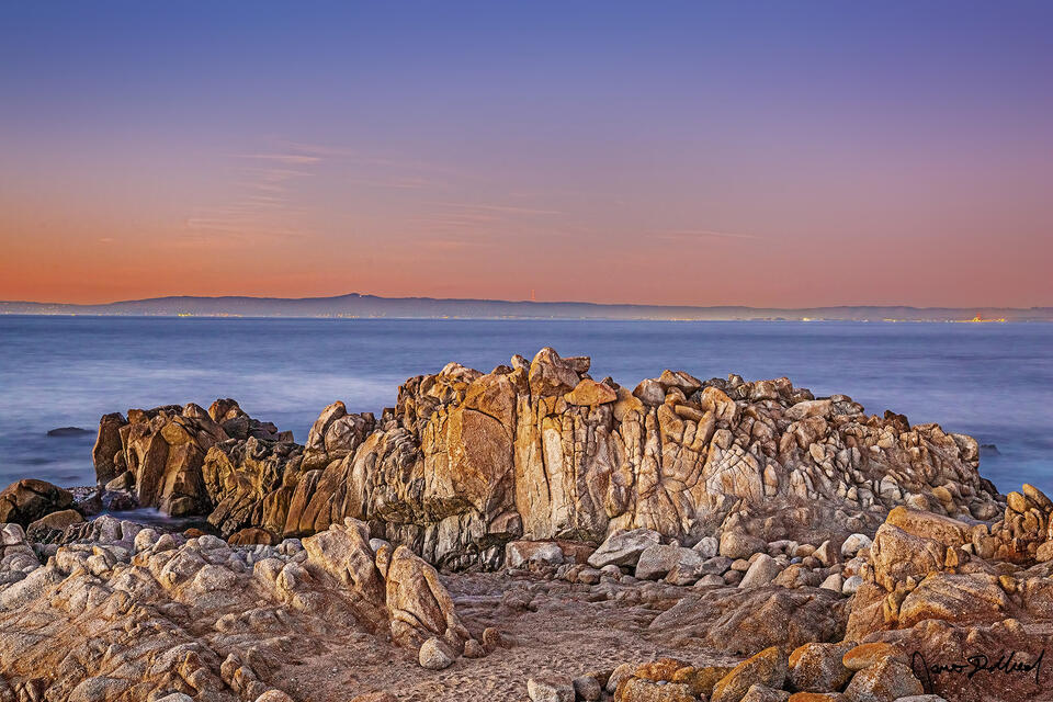 After sunset, the skies over the Pacific Ocean illuminate at dusk as civil twilight fades. Seagulls use a rocky outcrop just offshore for nesting and rest