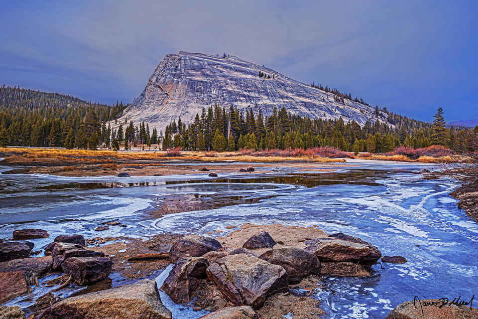 The Tuolumne River freezes over after a long cold spell as the first storm of winter nears. Lembert Dome reflects in the small patches of unfrozen water.