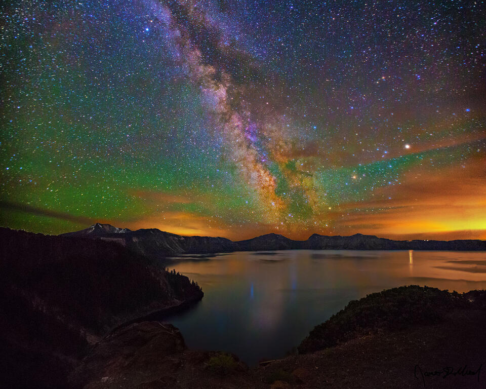 The sky illuminates green from diffuse aurora as the Milky Way core rises over Cleatwood Cove, reflecting in Crater Lake at Crater Lake National Park.