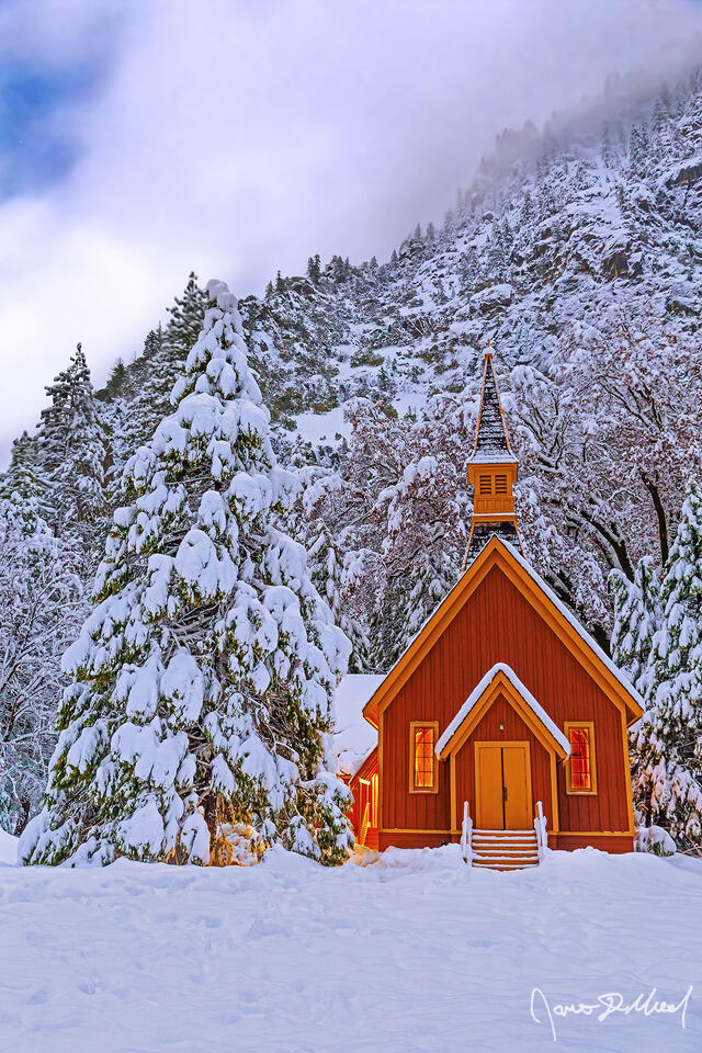 Snow blankets the Yosemite Valley Chapel, the surrounding trees and meadow at night as fog clings to the snow-covered granite cliffs rising 3000 feet above.