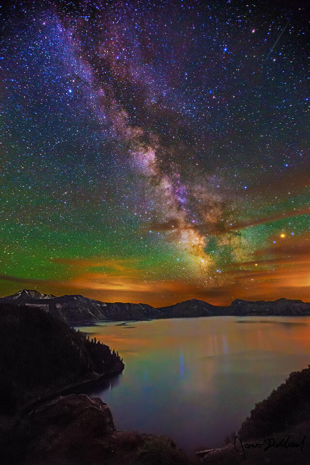 The Milky Way core rises above Cleetwood Cove and Crater Lake as green aurora induced airglow stretches across the sky.