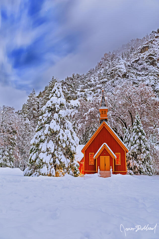 Snow blankets the Yosemite Valley Chapel, the surrounding trees and meadow at night as fog clings to the snow-covered granite cliffs rising 3000 feet above