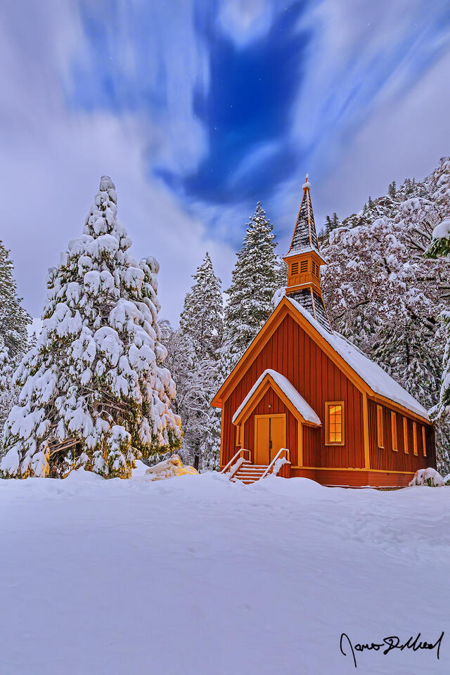 Snow blankets the Yosemite Valley Chapel, the surrounding trees and meadow at night as fog clings to the snow-covered granite cliffs rising 3000 feet above