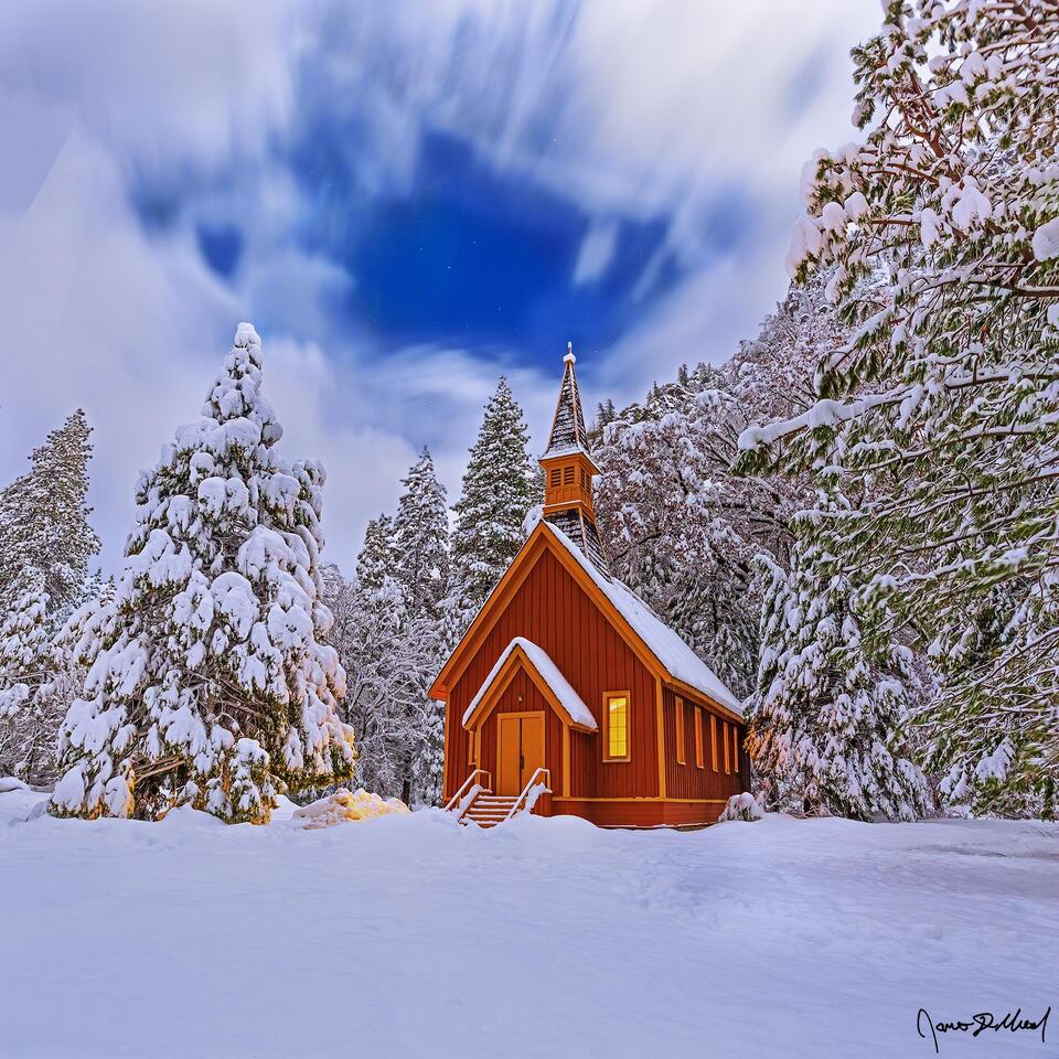 Snow blankets the Yosemite Valley Chapel, the surrounding trees and meadow at night as fog clings to the snow-covered granite cliffs rising 3000 feet above.