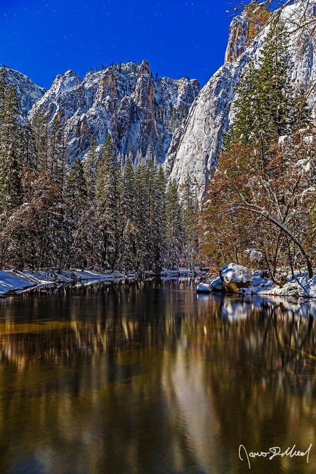 Moonlight illuminates snow covered Cathedral Rocks above Yosemite Valley in the winter as the still, clear waters of the Merced River faintly reflect the trees.