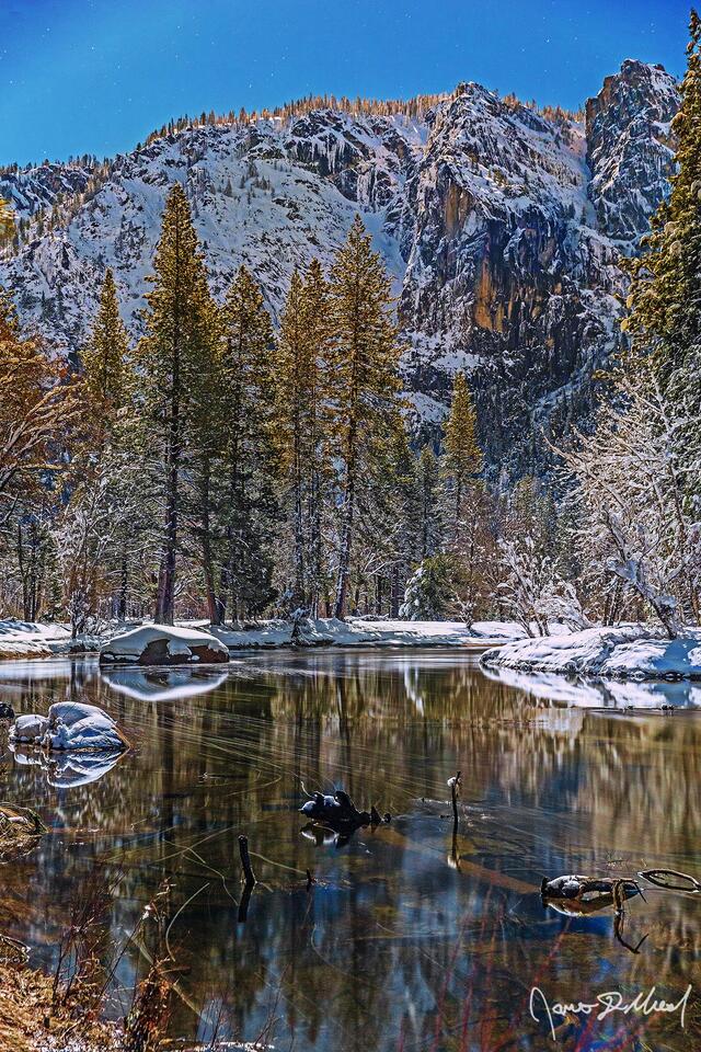 Moonlight illuminates the snow covered granite cliffs above Yosemite Valley in the winter as the still, clear waters of the Merced River reflect the trees .
