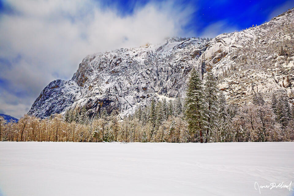 Deep snow covers Cooks Meadow in Yosemite National Park after a winter snow storm, as Moonlight illuminates the granite snow-covered cliffs of Yosemite Valley.