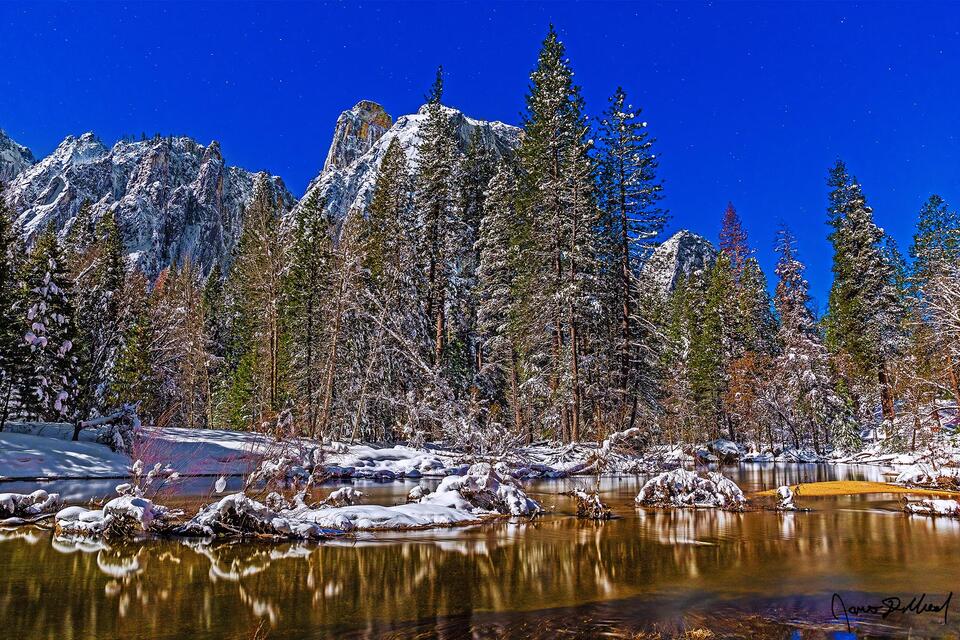Yosemite Valley lay covered in a blanket of snow in the winter as moonlight illuminates snow covered Cathedral Rocks above the valley and the Merced River.