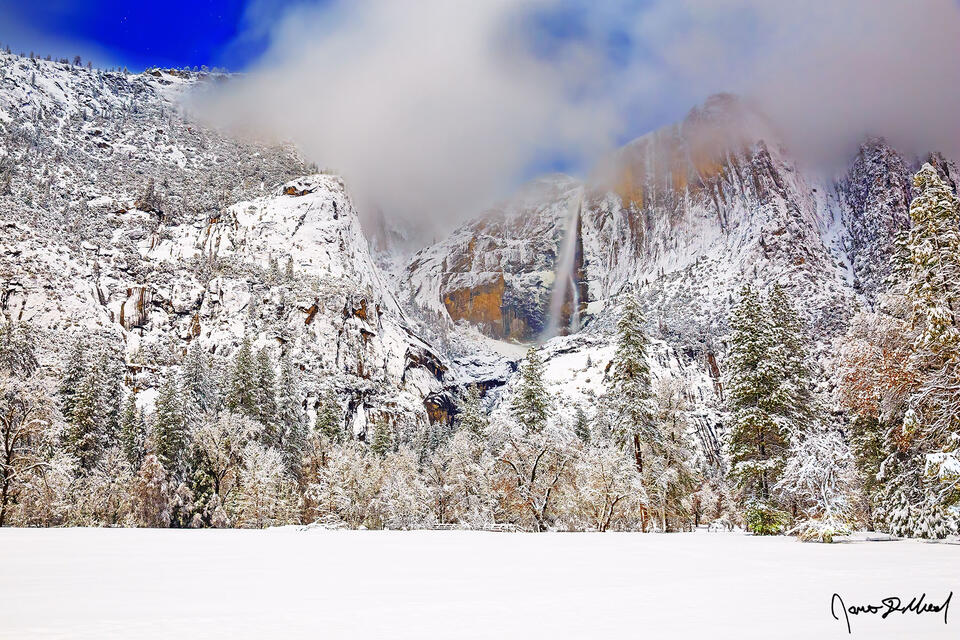 Moonlight illuminates the clouds in the sky, Yosemite Falls and the granite cliffs above Yosemite Valley as deep winter snow blankets Cooks Meadow.