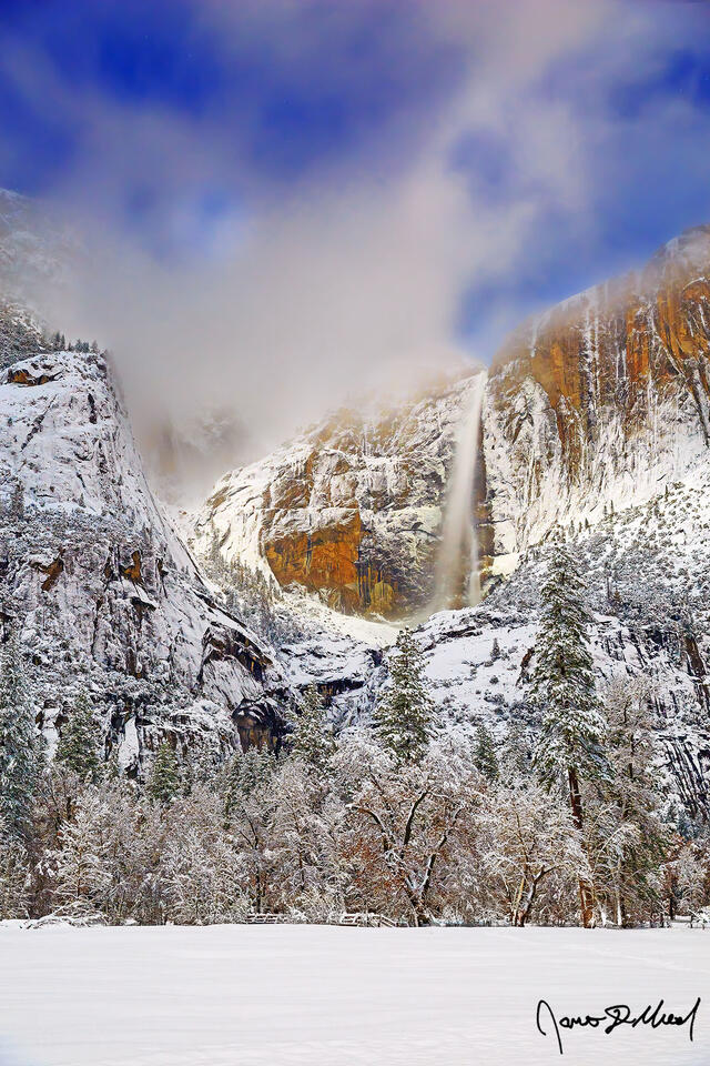 Moonlight illuminates the clouds in the night sky, Yosemite Falls and the granite cliffs above Yosemite Valley as deep winter snow blankets Cooks Meadow