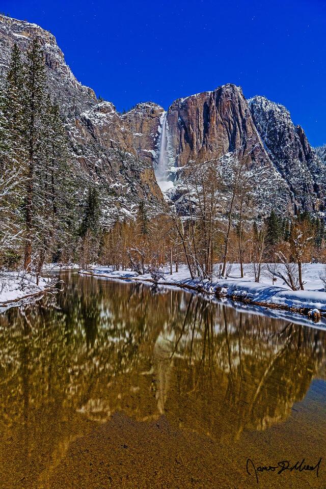 Moonlight illuminates the cliffs above snow covered Yosemite Valley as Yosemite Falls tumbles over the cliff as the scene reflects in the Merced River below.