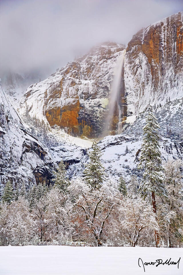 Moonlight shining through cloud breaks in the night sky illuminates Yosemite Falls and the snow covered granite cliffs above snow covered Yosemite Valley.