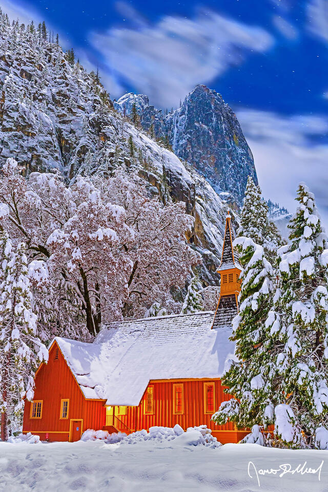 Snow blankets the Yosemite Valley Chapel, the surrounding trees and meadow at night as fog clings to the snow-covered granite cliffs rising 3000 feet above