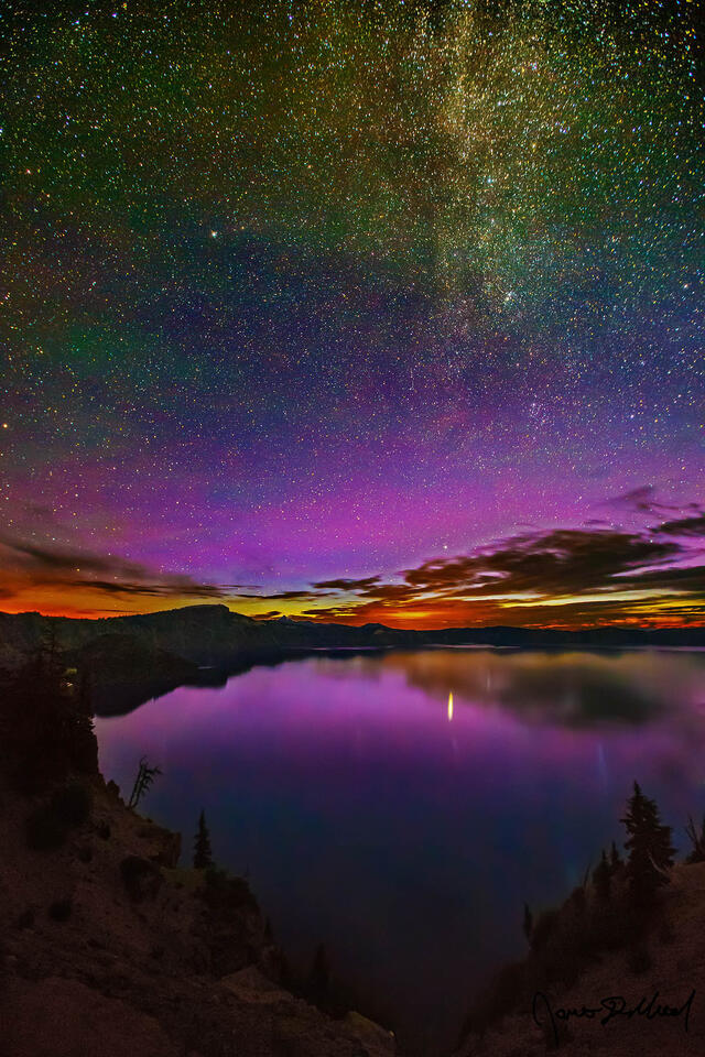 Purples of the Aurora Borealis reflect off of Crater Lake as light from the rising sun still far below the horizon can be seen just above the caldera rim.
