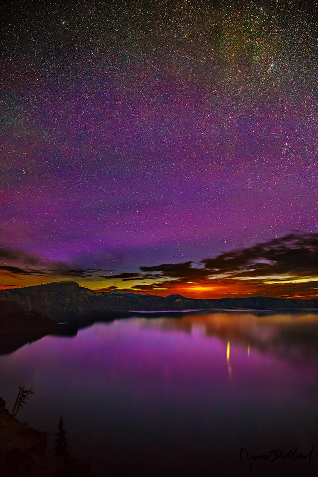 The aurora borealis reflect off Crater Lake as light from the rising sun still far below the horizon can be seen just above the caldera rim.