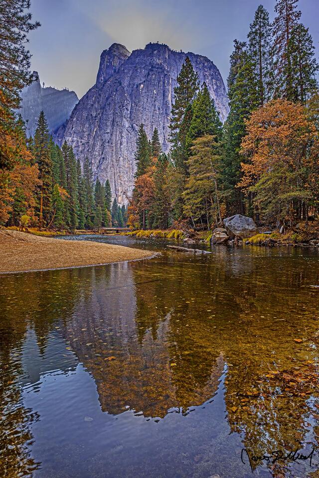 Light Beams shine from behind Cathedral Rocks and Spires as colors of fall line the banks of the Merced River in Yosemite Valley.