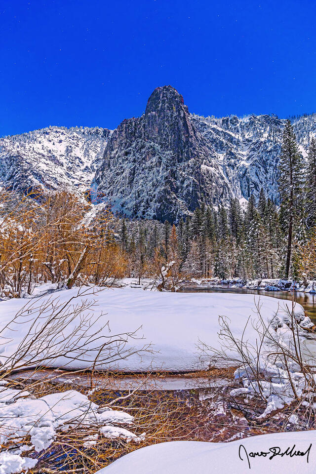 The moon illuminates the night skies above Sentinel Rock and the Merced River as snow blankets the Yosemite valley floor and granite cliffs rising above.