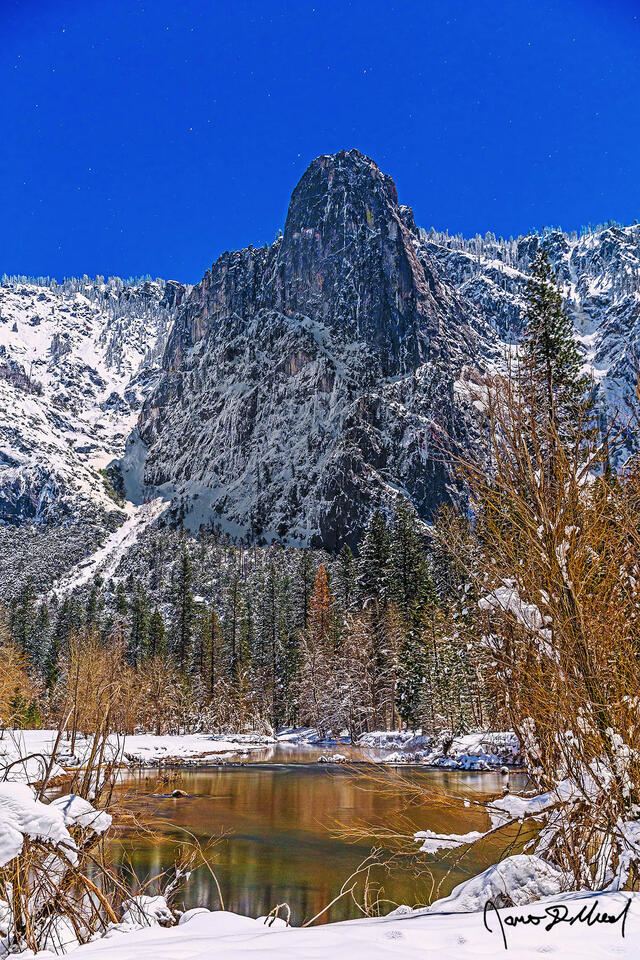The moon illuminates the night skies above Sentinel Rock and the Merced River as snow blankets the Yosemite valley floor and granite cliffs rising above.
