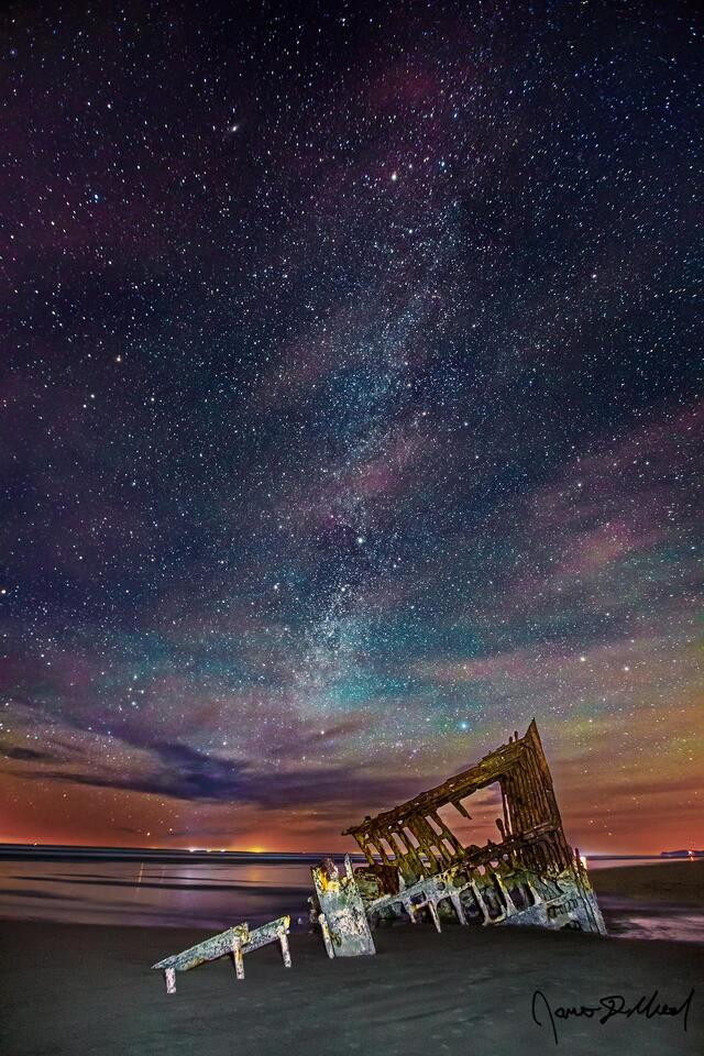 The winter Milky Way rises over the Wreck of the Peter Iredale and the Pacific Ocean at night on the Oregon Coast as airglow illuminates the night sky.