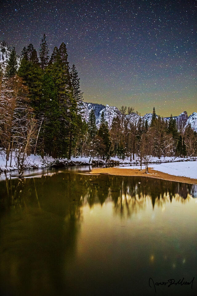 Snow Yosemite Valley reflects in the still, cold waters of the Merced River during winter as the stars in the night sky shine brightly overhead