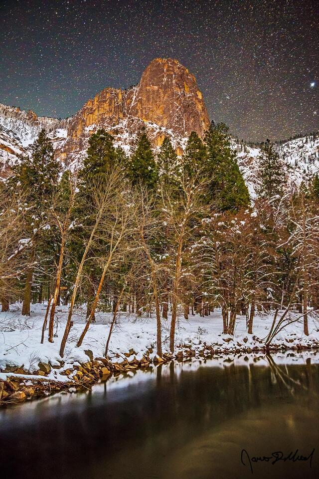 Stars shine bright in the night skies above Sentinel Rock and the Merced River during winter as snow blankets the Yosemite valley floor and granite cliffs above