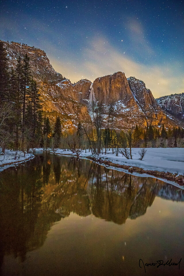 The rising moon in the east illuminates the granite cliffs of Yosemite Valley. Yosemite Falls tumbles over the cliffs reflecting in the Merced River below.