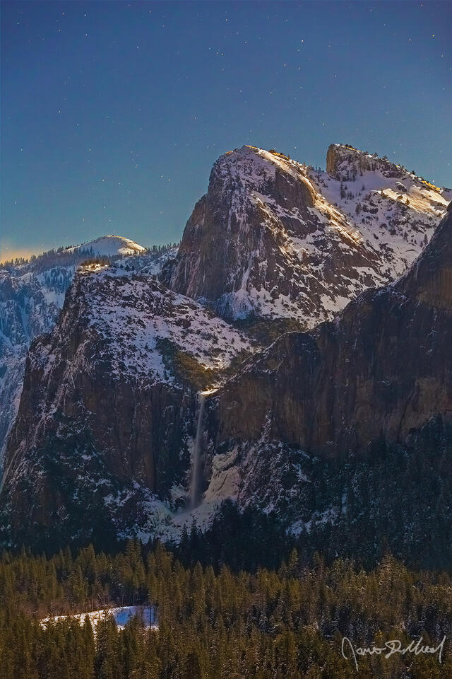 Moonlight illuminates snow covered Cathedral Rocks and Cathedral Spires as Bridalveil Fall flows over 620 feet the cliffs towards snow covered Yosemite Valley.