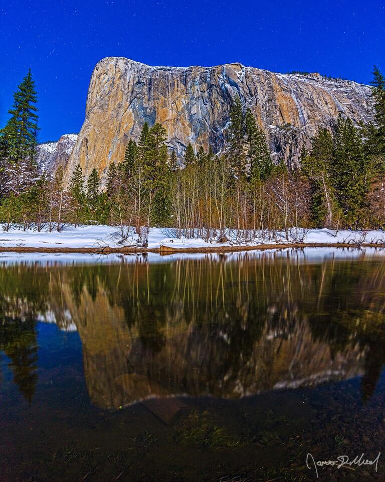 The moon illuminates El Capitan in the winter as the granite monolith reflects in the cold, clear waters of the Merced River. and snow covered Yosemite Valley.