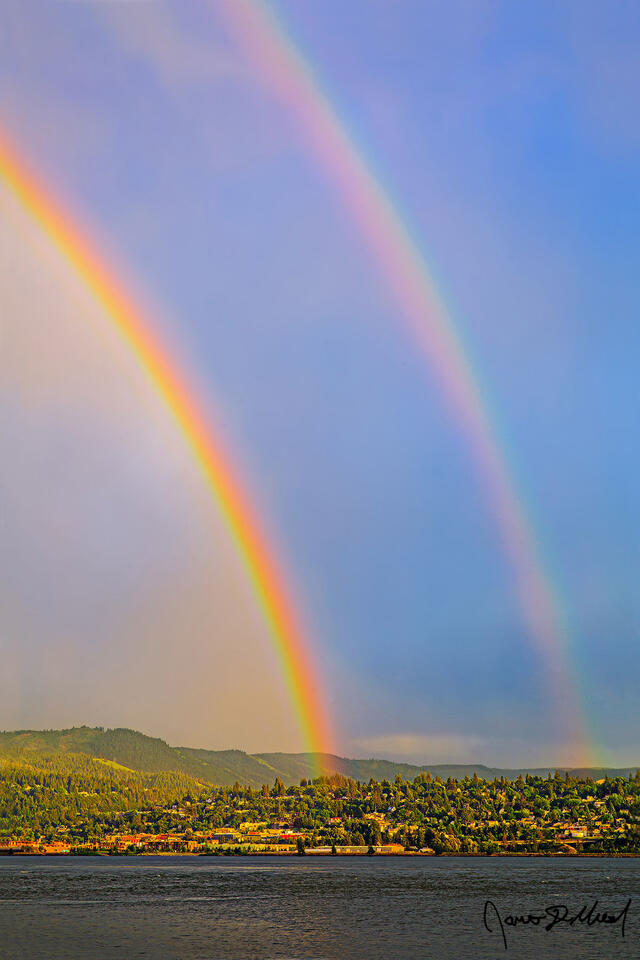 A double rainbow forms over Hood River and the Columbia River at the beginning of the golden hour as soft pastel colors illuminate the clouds in the sky.