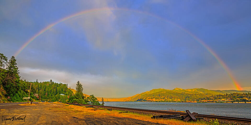 A rainbow forms over over Hood River and the Columbia River at the beginning of the golden hour as soft pastel colors illuminate the clouds in the sky.