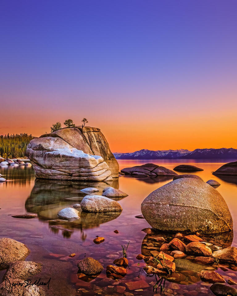Sunset brings soft pastel colors and a warm golden glow to the skies above Bonsai Rock, as the large granite boulders reflect in the waters of Lake Tahoe.