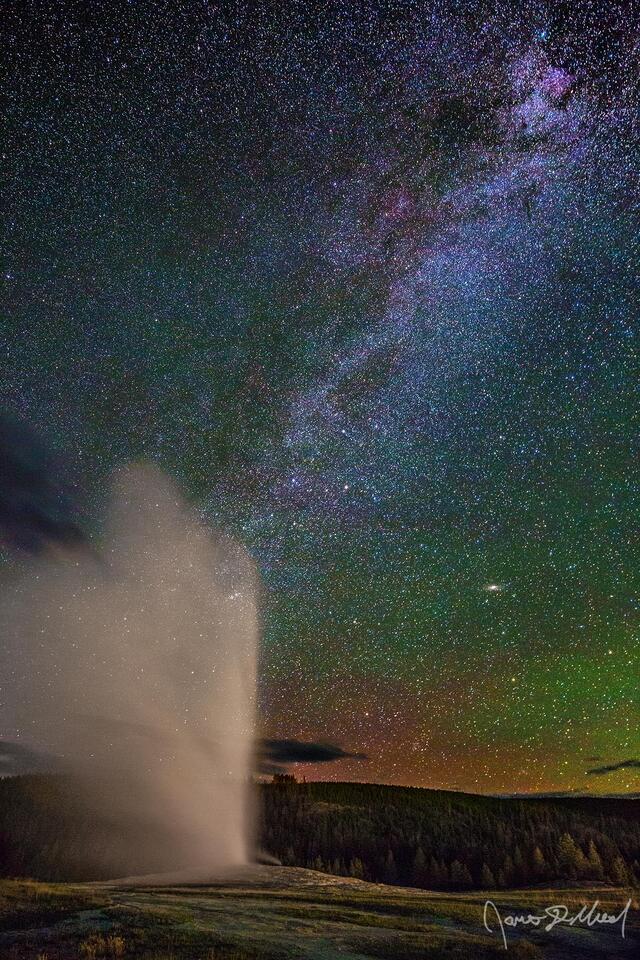 Old Faithful erupts as the Milky Way spiral arm rises into the sky while magenta from a faint Aurora Borealis begins to show on the horizon.