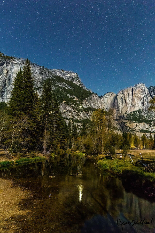 A climbers light reflects in the still waters of the Merced River as the stars illuminate the night sky above Yosemite Valley, the Three Brothers and Eagle Peak