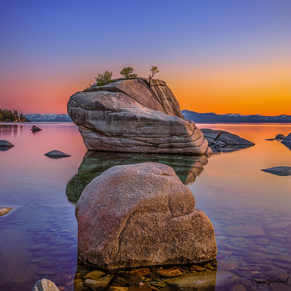 Sunset bathes the skies above Bonsai Rock and Lake Tahoe in soft, golden pastel light as the colors reflect in the crystal clear mirror like waters of the lake 
