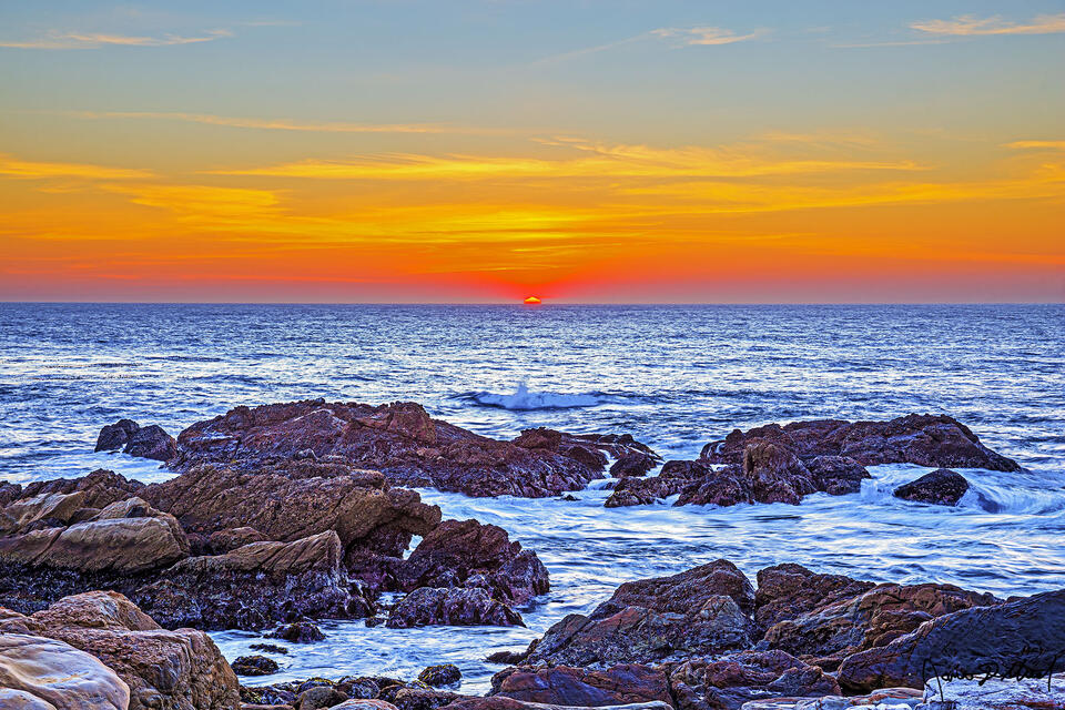 The sun dips below the horizon at sunset as waves from the Pacific Ocean crash onto the rocks near the rugged shoreline.